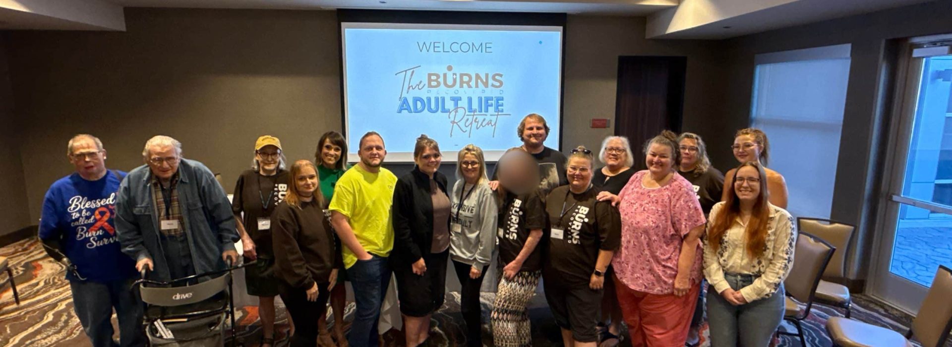 A group of people standing in a conference room in front of a projection screen that reads “Welcome The Burn Adult Life Retreat.”