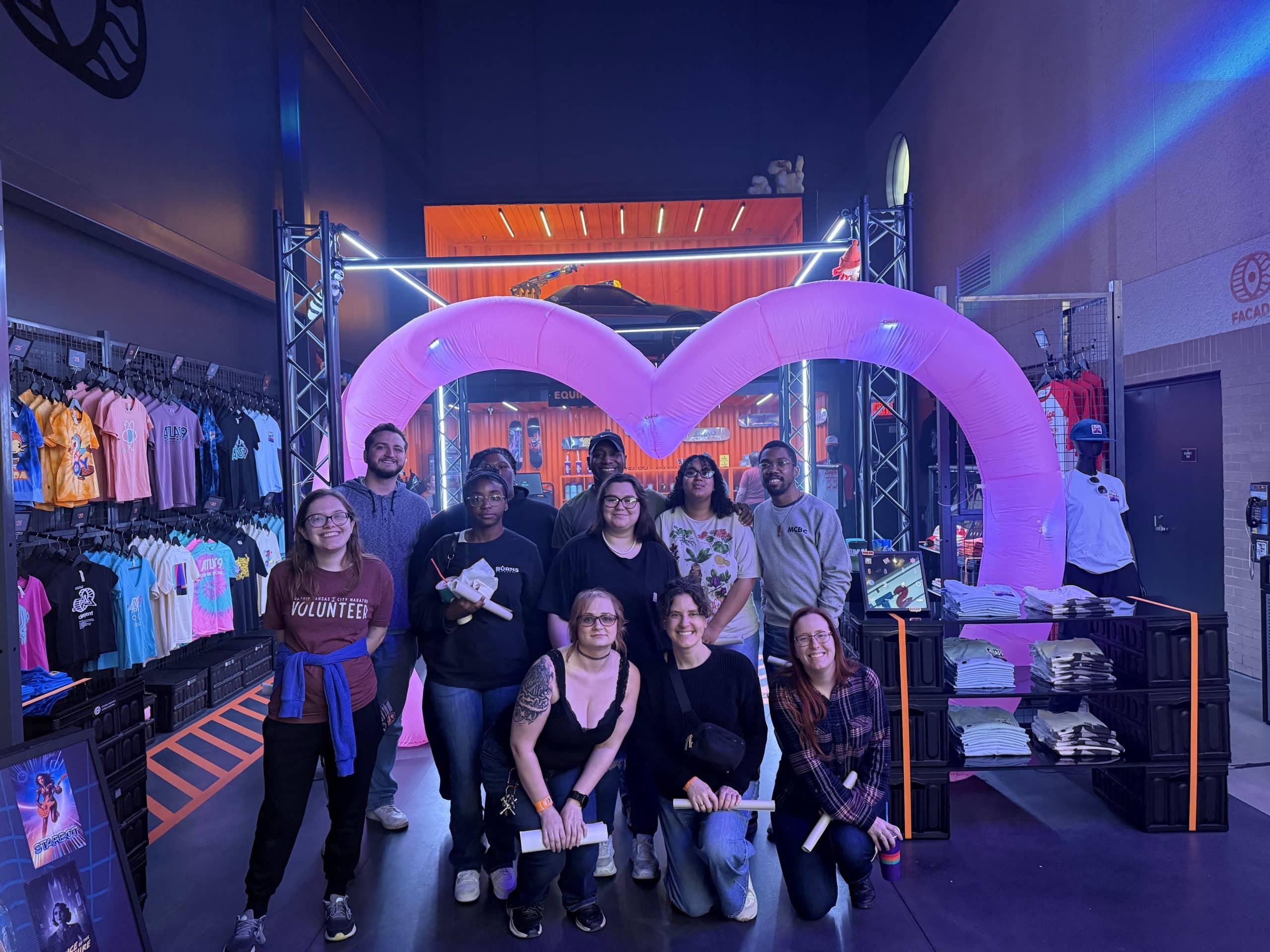 A group of twelve people posing in front of a large, pink, heart-shaped inflatable structure inside a retail store.