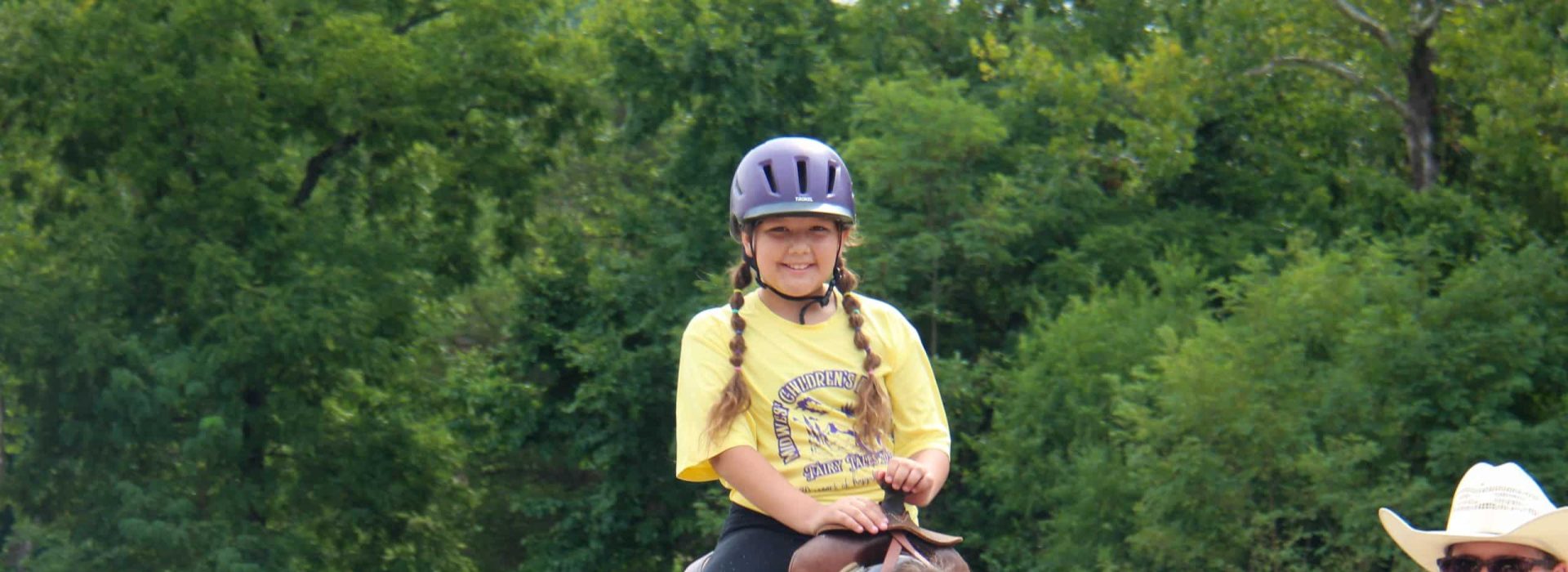 A girl with braided hair and a purple helmet riding a brown horse in an outdoor arena, with a person wearing a cowboy hat walking alongside.