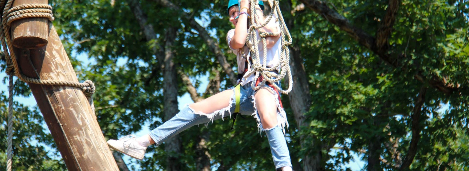 A person wearing a helmet and climbing harness swings from a rope ladder on a ropes course.