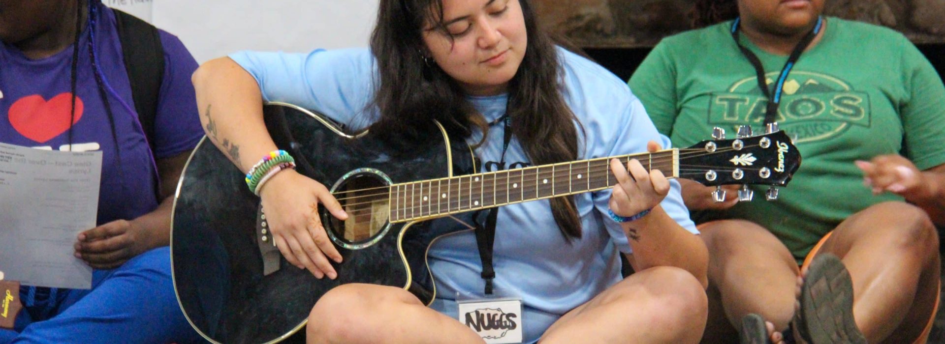 Three people sitting on the floor; the center person plays an acoustic guitar and wears a flower crown.