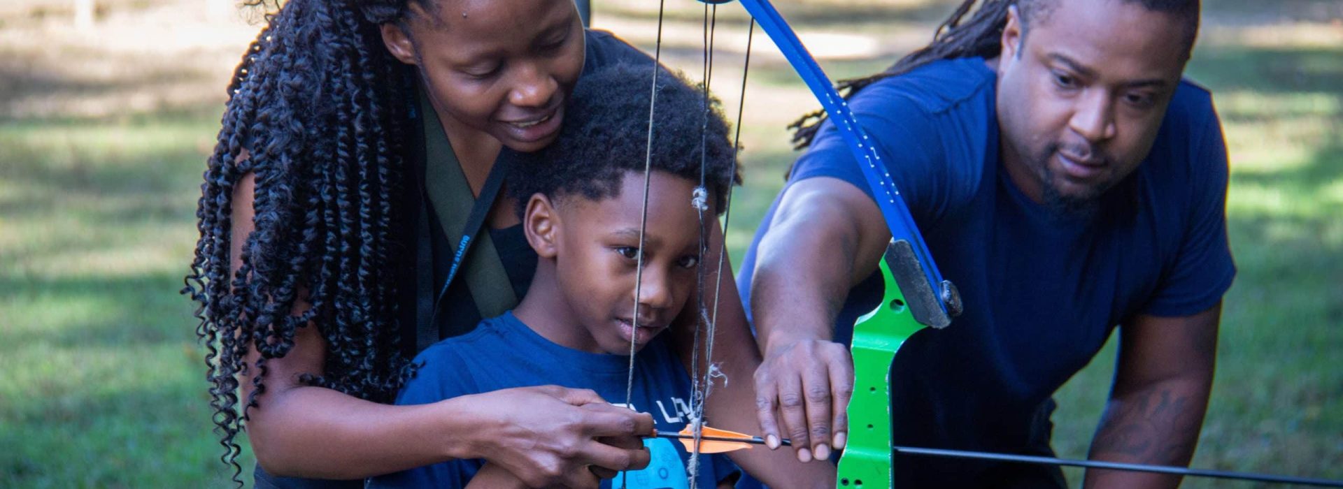 A woman and a man help a young boy hold a compound bow and arrow outdoors.