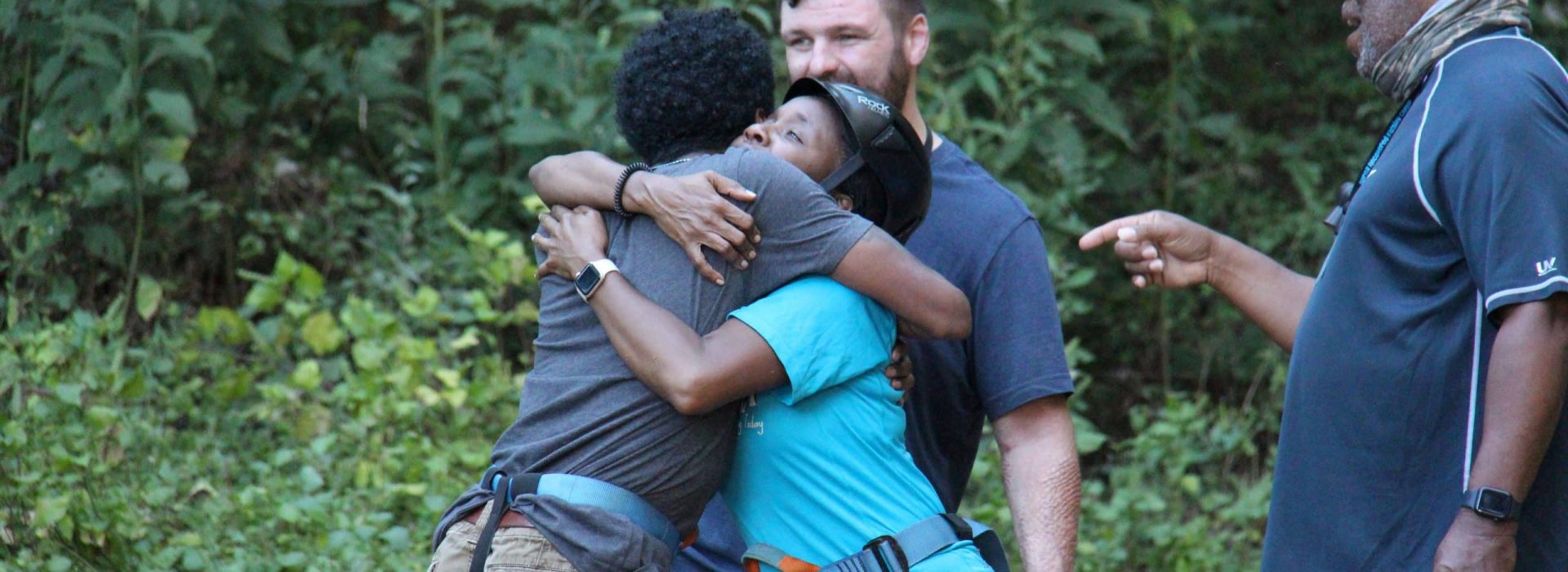Two people in climbing harnesses hug each other as a man watches nearby.