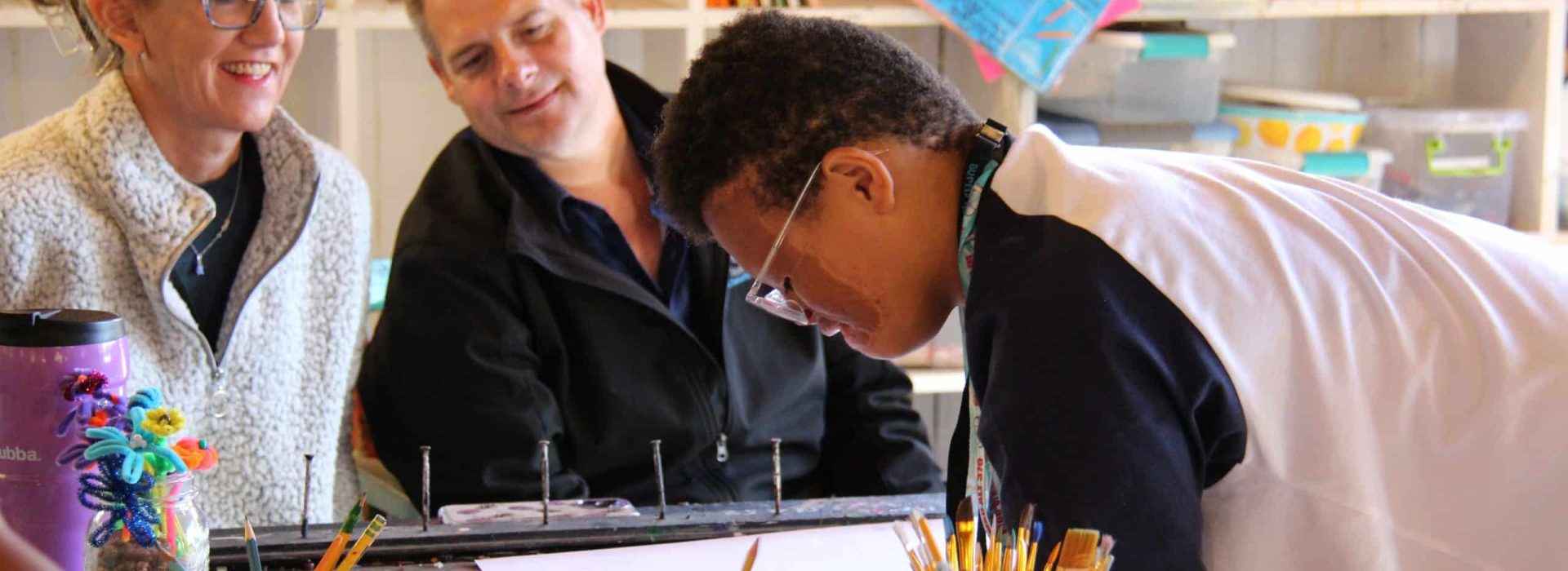 A young person draws on paper while a woman and a man watch from across a craft table.
